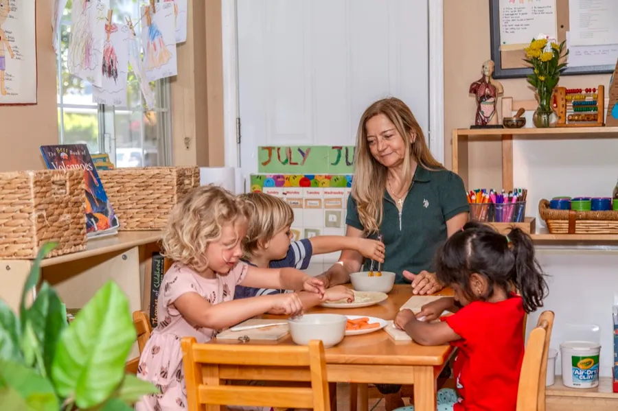 A teacher and preschoolers preparing snacks together around a classroom table