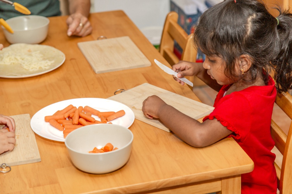 A preschooler practicing knife skills by cutting carrots at a wooden table