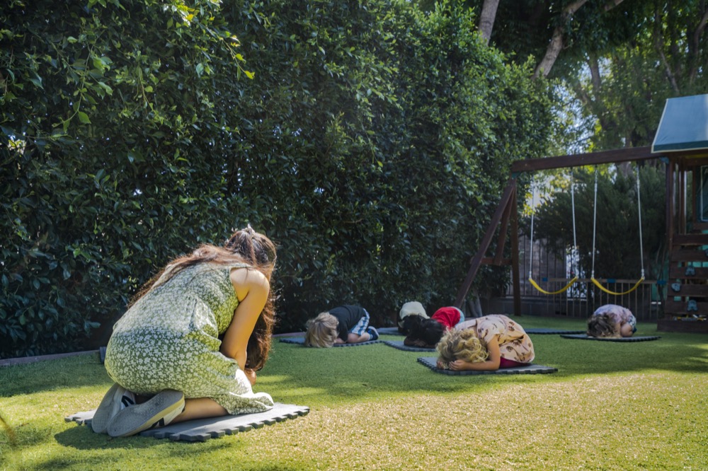 A teacher and children practicing yoga in child’s pose on mats in the outdoor play area