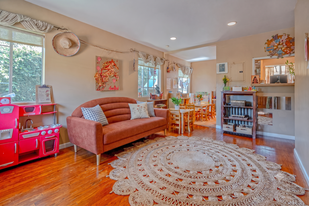 A warm preschool playroom with a terracotta sofa, child-sized tables, and a red play kitchen on hardwood floors